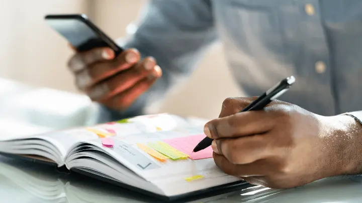 Man writing into notebook while holding a phone.